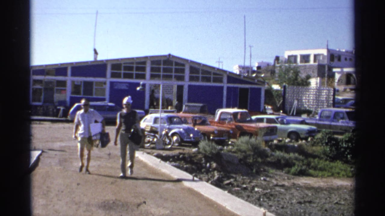Two adults continue walking past parked vehicles outside a blue building.