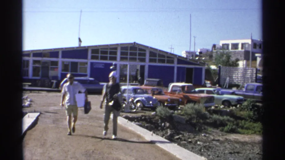 Two adults carrying items approach the camera on a sunlit dirt path in Guaymas, with 1970s cars and a blue building behind them.
