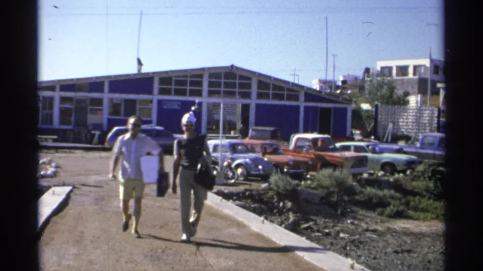 Two adults pass closer to the camera in Guaymas, Mexico, carrying bags with a row of classic cars and a blue building in the background.