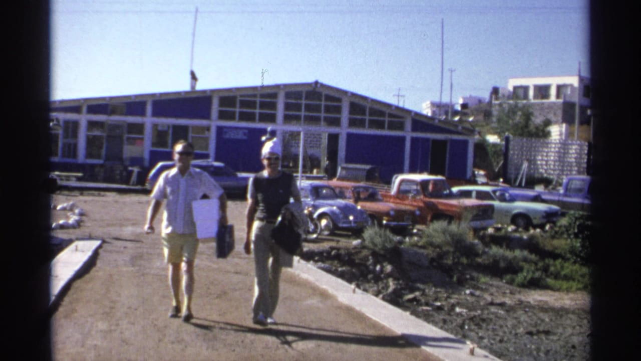 Two adults walk along a marina path near parked cars and a blue-and-white building.