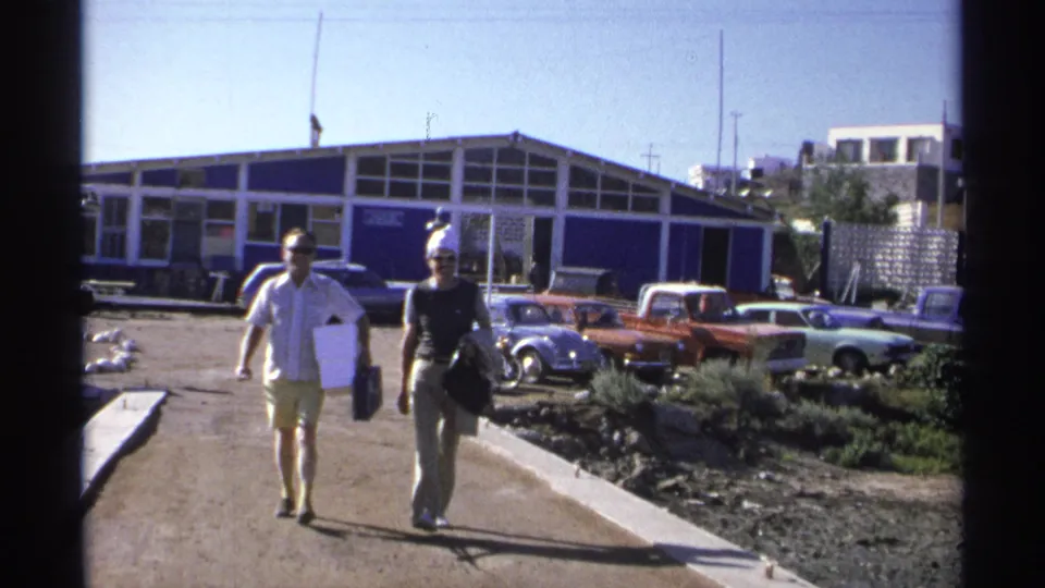 In 1975 Guaymas, Mexico home-movie footage, a couple carrying bags walks near a marina building and parked cars.