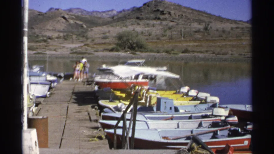 People stand near small boats on a Guaymas marina dock, framed by calm water and dry hills in 1975 8mm footage.