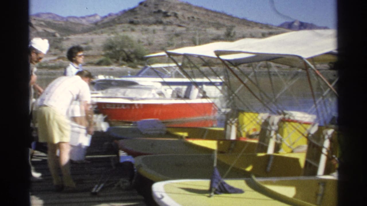 Adults stand and bend near moored boats on a sunny dock with a cooler nearby.