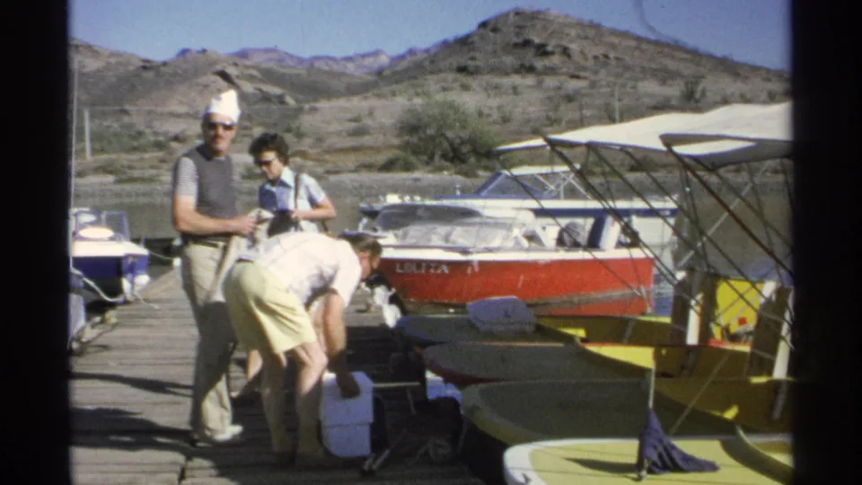 Three adults stand or bend near boats on a 1975 Guaymas dock, with a red motorboat, yellow canopies, and hills behind them.