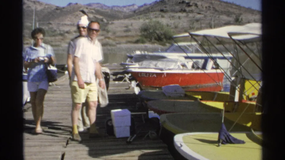 Adults in summer clothing walk along a Guaymas dock in 1975, passing yellow boats, a cooler, a red motorboat, water, and arid hills.