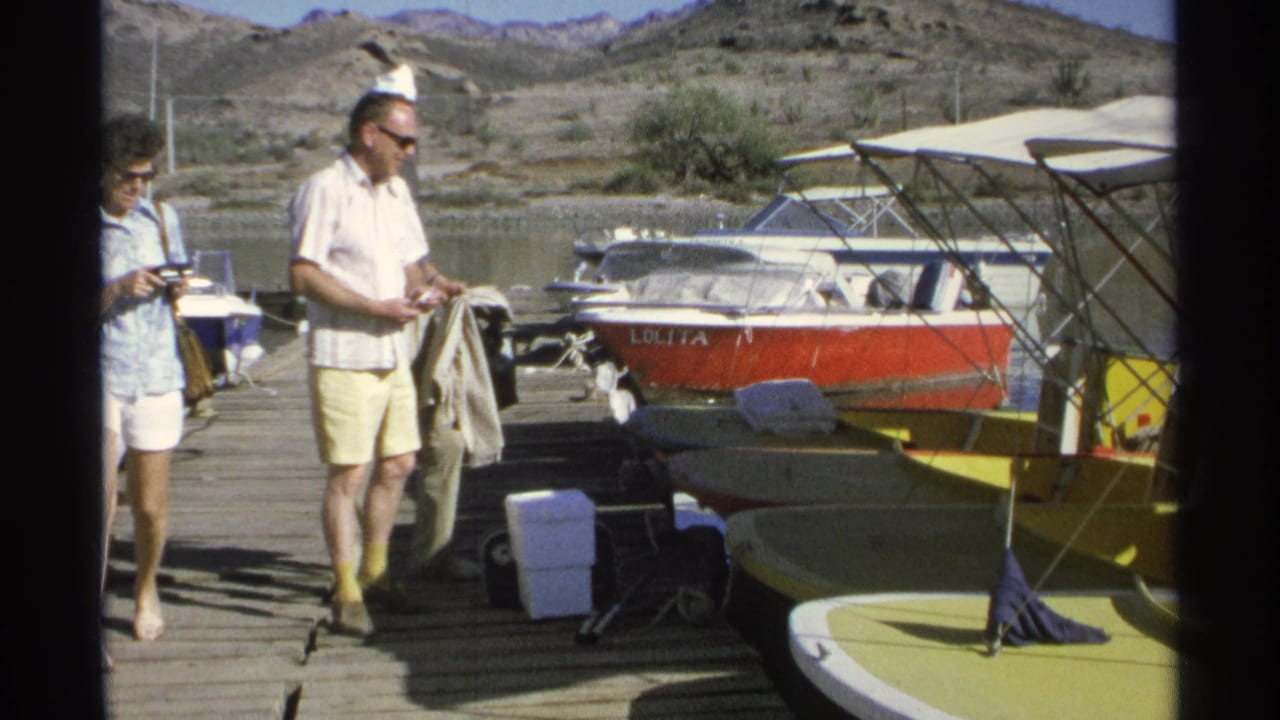 Two adults walk along a wooden dock beside small moored boats and dry hills.