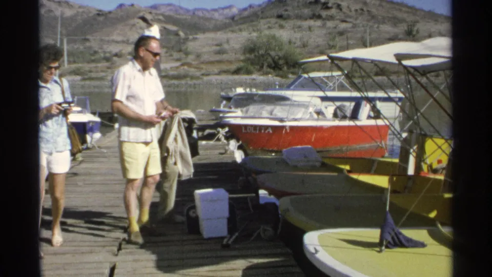 1975 8mm home-movie frame from Guaymas, Mexico, showing two adults on a dock near small boats and arid hills.