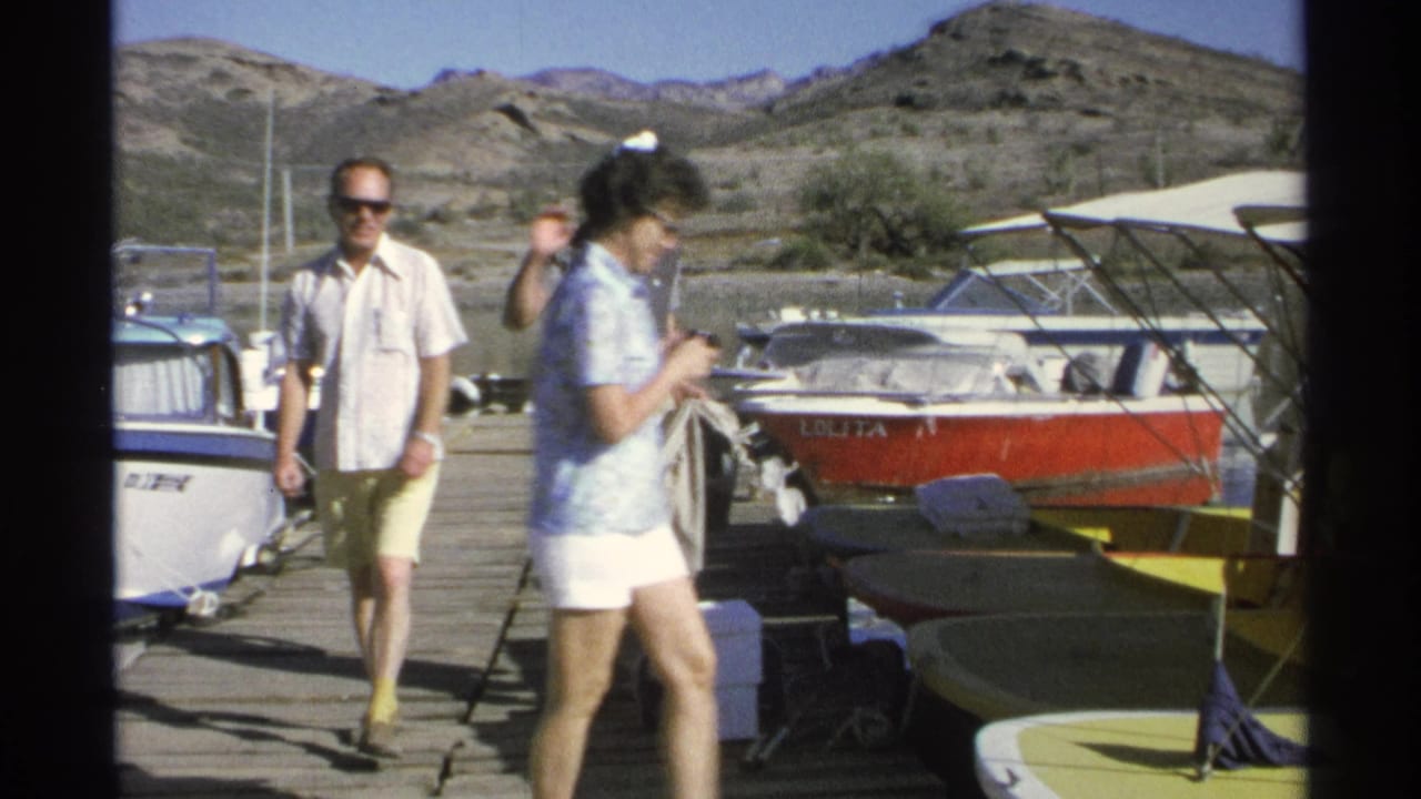 A woman crosses the dock while a man stands near boats, with a red boat behind them.