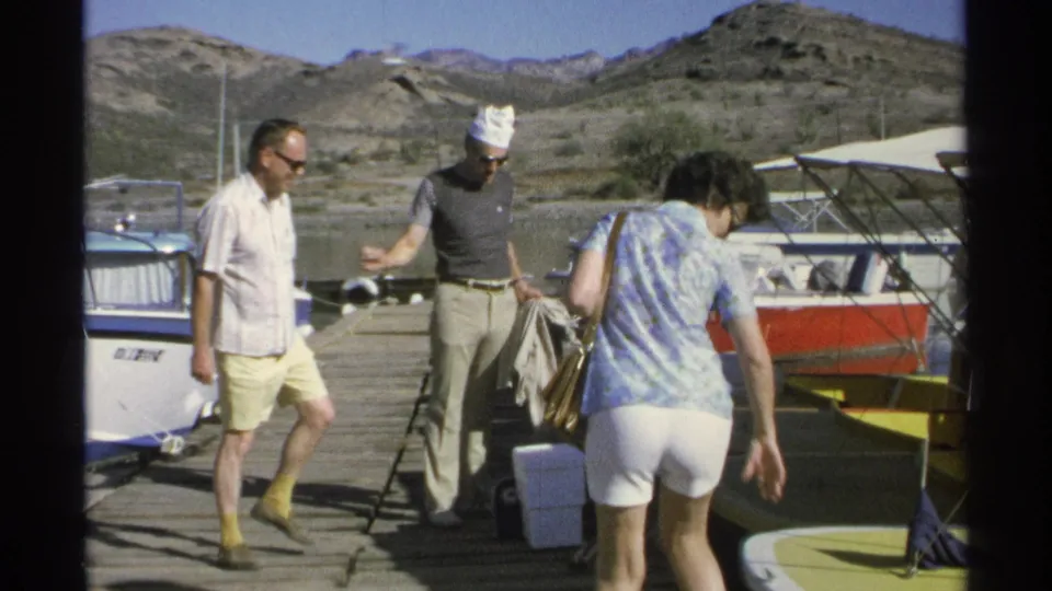 1975 8mm frame in Guaymas showing three adults around luggage on a dock beside moored boats.