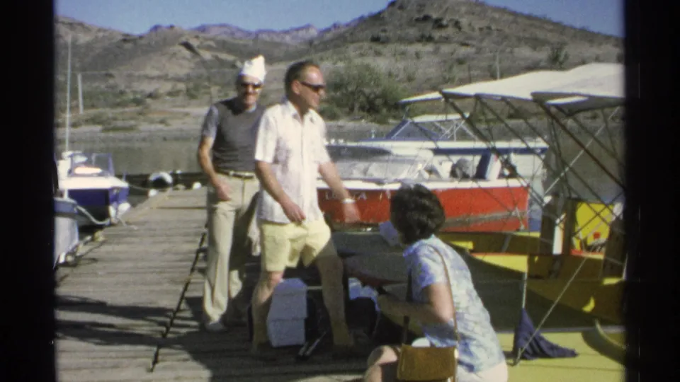 1975 Guaymas home-movie frame with two men on the dock and a woman low beside a yellow boat near the water.