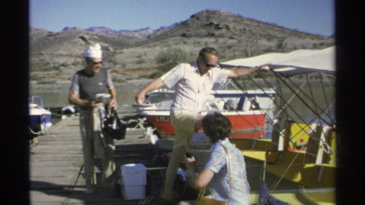 Two men and a seated person near small boats on a wooden dock with rocky hills behind.