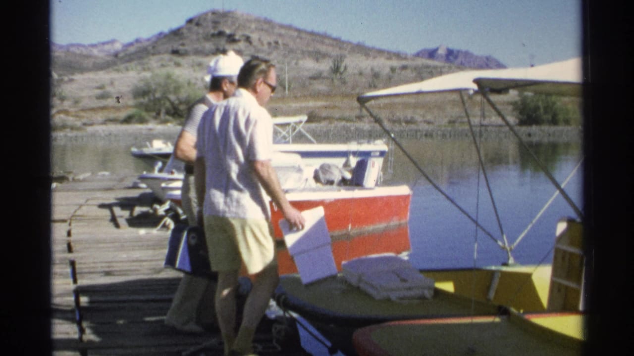 Two men walk along a dock beside red and yellow boats with coolers and water nearby.