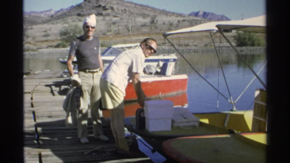 A man in sunglasses bends near supplies while another man in a white cap stands by the docked boats in Guaymas, Mexico.