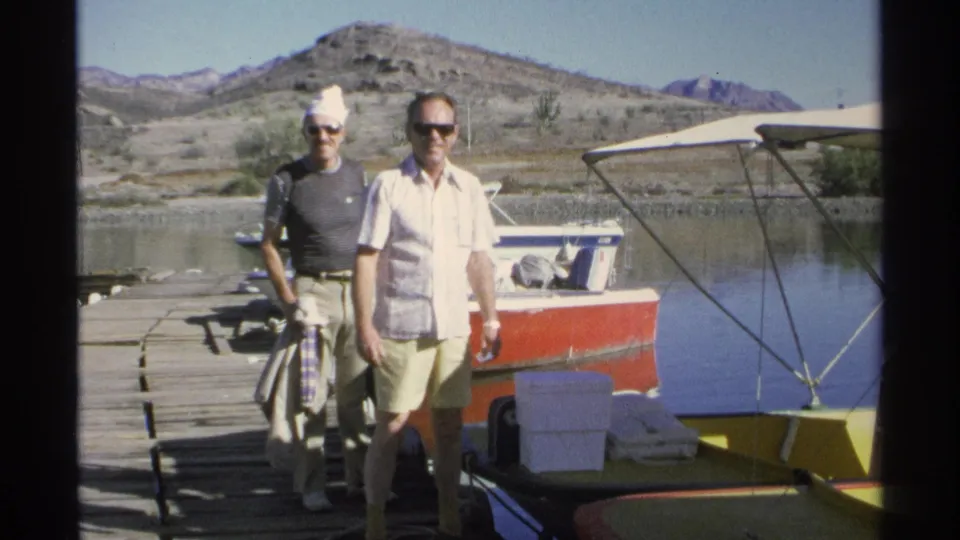 Two adult men on a sunny Guaymas dock face the camera near loaded small boats, coolers, water, and rocky hills.