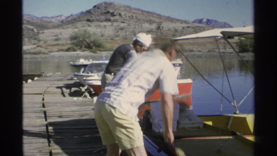 One man bends forward at the dock while another stands nearby with gear beside small boats in the 1975 Guaymas home-movie scene.