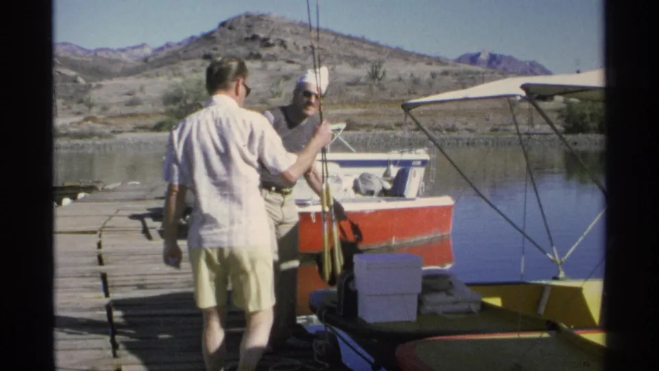Two adult men confer near a docked motorboat as fishing rods and boating gear appear in the 1975 Guaymas, Mexico footage.