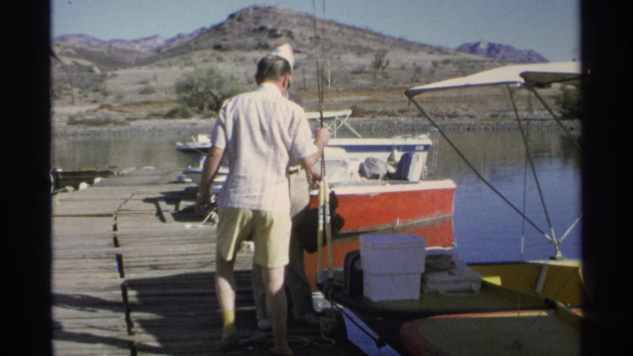 Two men stand on a wooden dock beside boats with fishing rods and gear.