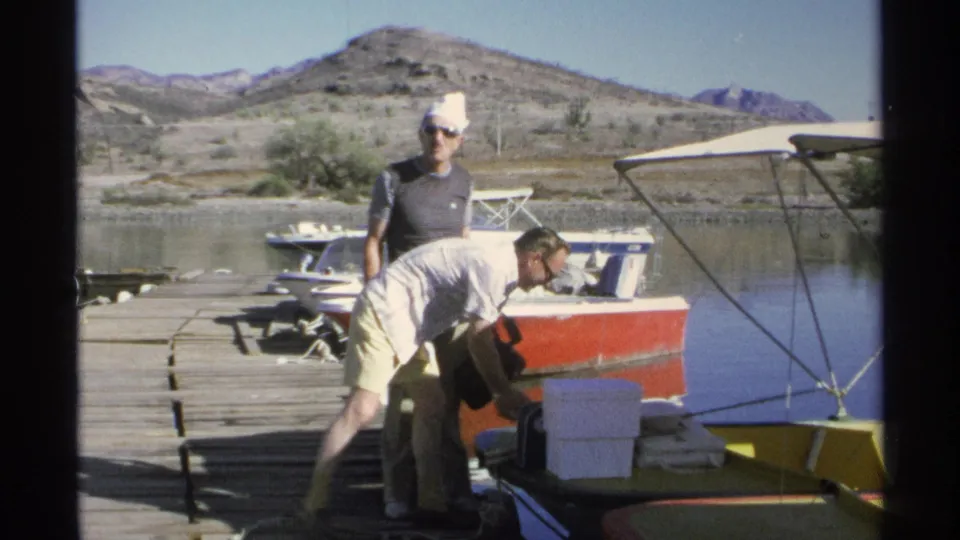 1975 Guaymas home-movie still of a man leaning over fishing gear on a dock with boats, water and desert hills in view.