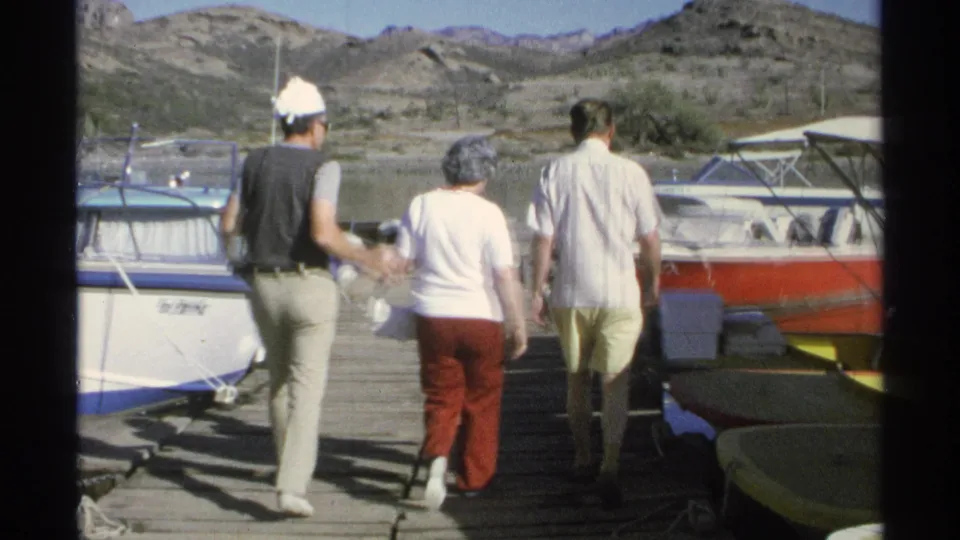 1975 Guaymas frame of two men and a woman walking away on a wooden dock between moored small boats.
