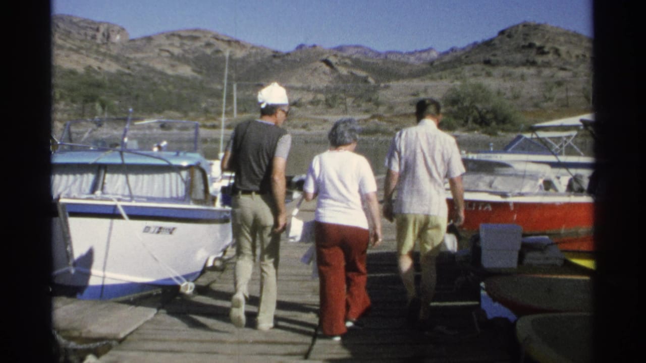 Three adults walk along a wooden dock between moored boats with dry hills behind them.