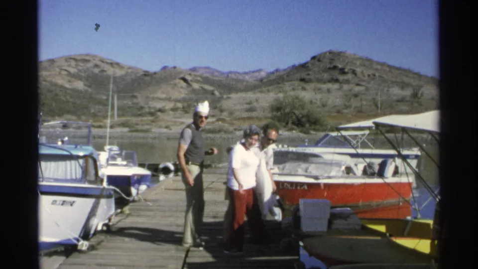On a 1975 Guaymas dock, the men and older woman gather beside moored boats before boarding or moving toward a vessel.