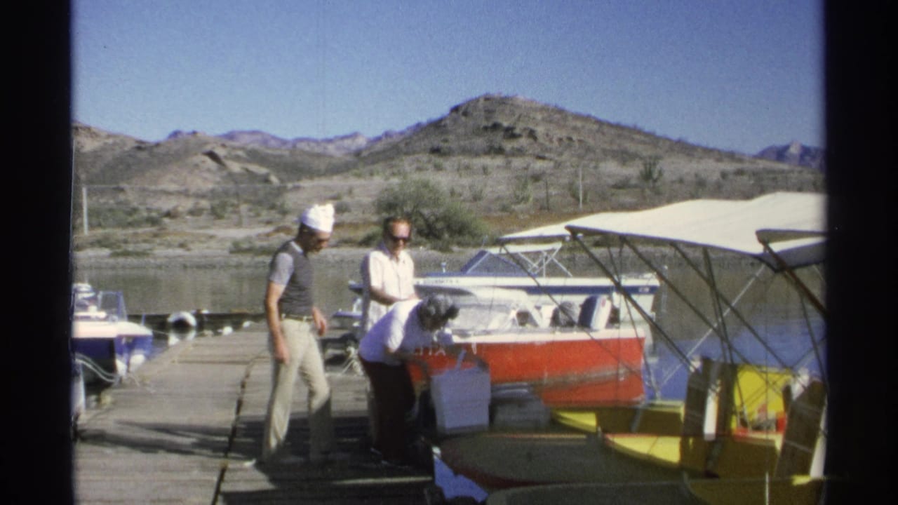 Three men stand by a docked boat with other small boats and rocky hills behind them.