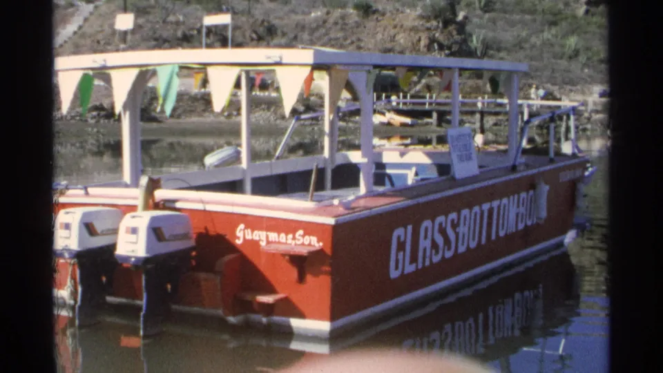 A red glass-bottom boat with a white canopy and colorful pennants sits on calm water in Guaymas, Mexico.