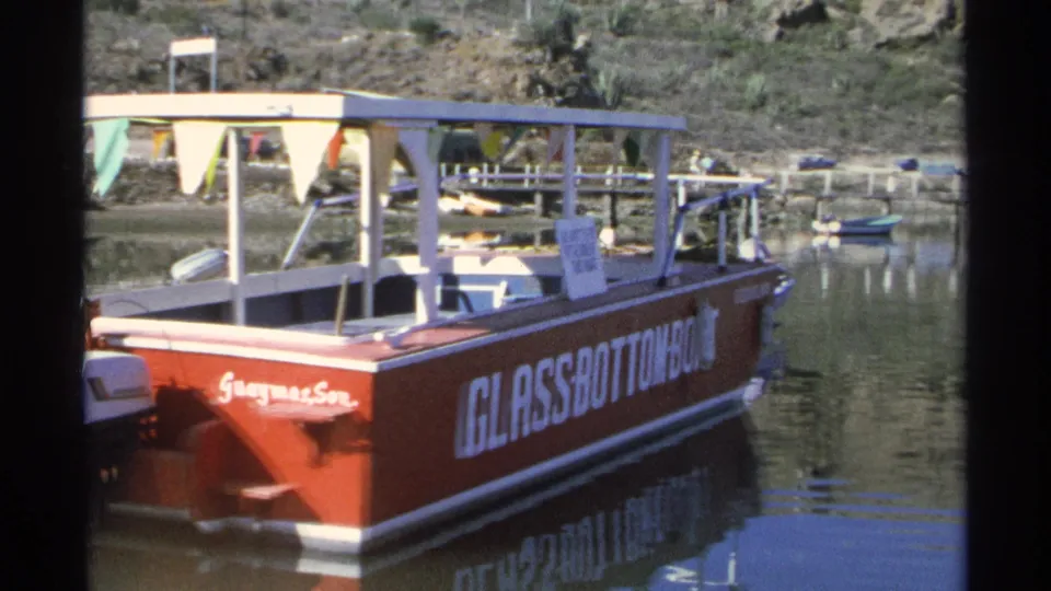 The red glass-bottom boat is seen from the side, with its canopy, flags, motors, and large lettering reflected on the water.