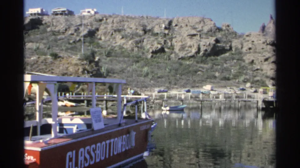 A docked glass-bottom boat appears below rocky hills in Guaymas, Mexico, with small boats and shoreline structures nearby.