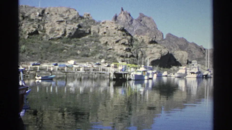 Wide 1975 Guaymas marina view with small boats tied near docks, glassy reflections, rugged hills, and clear sky.
