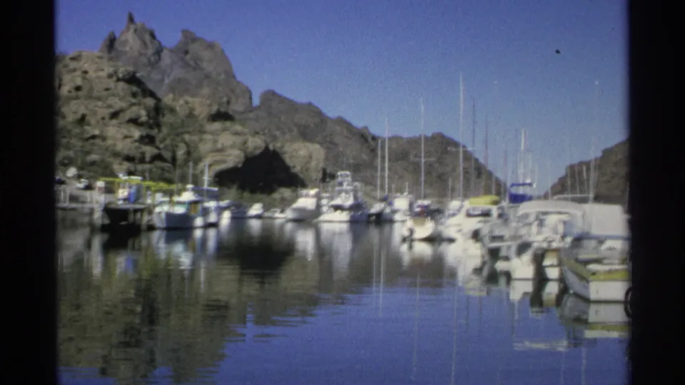 1975 home-movie frame of a quiet Guaymas marina, showing docked boats, reflective water, and rugged hills.