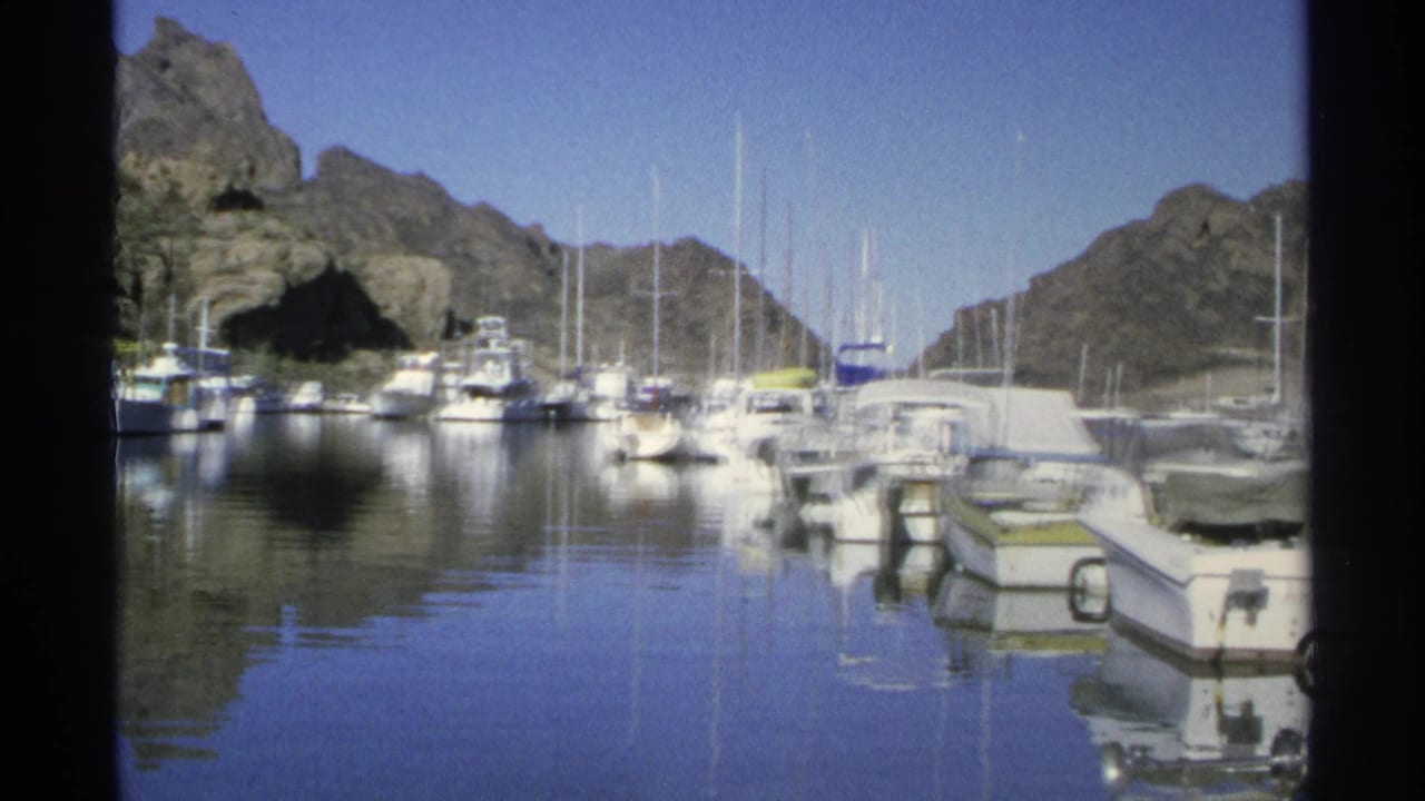 1975 8mm view of boats moored in Guaymas harbor, with mast reflections on still water and rocky mountains in the background.