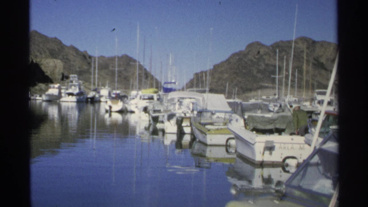 Harbor boats and sailboat masts reflected in still water near rocky hills.