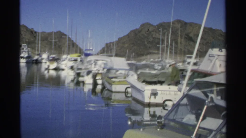 1975 Guaymas harbor frame with a man in green near a moored boat, surrounded by sailboats, docks, still water, and mountains.