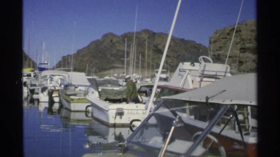 A man in a hat and green jacket appears on a foreground boat in the 1975 Guaymas marina, with boats, masts, and rocky mountains behind him.