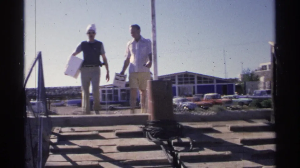 Two adult men in casual summer clothing walk on a Guaymas marina dock, one carrying a box, with buildings and parked cars behind them.