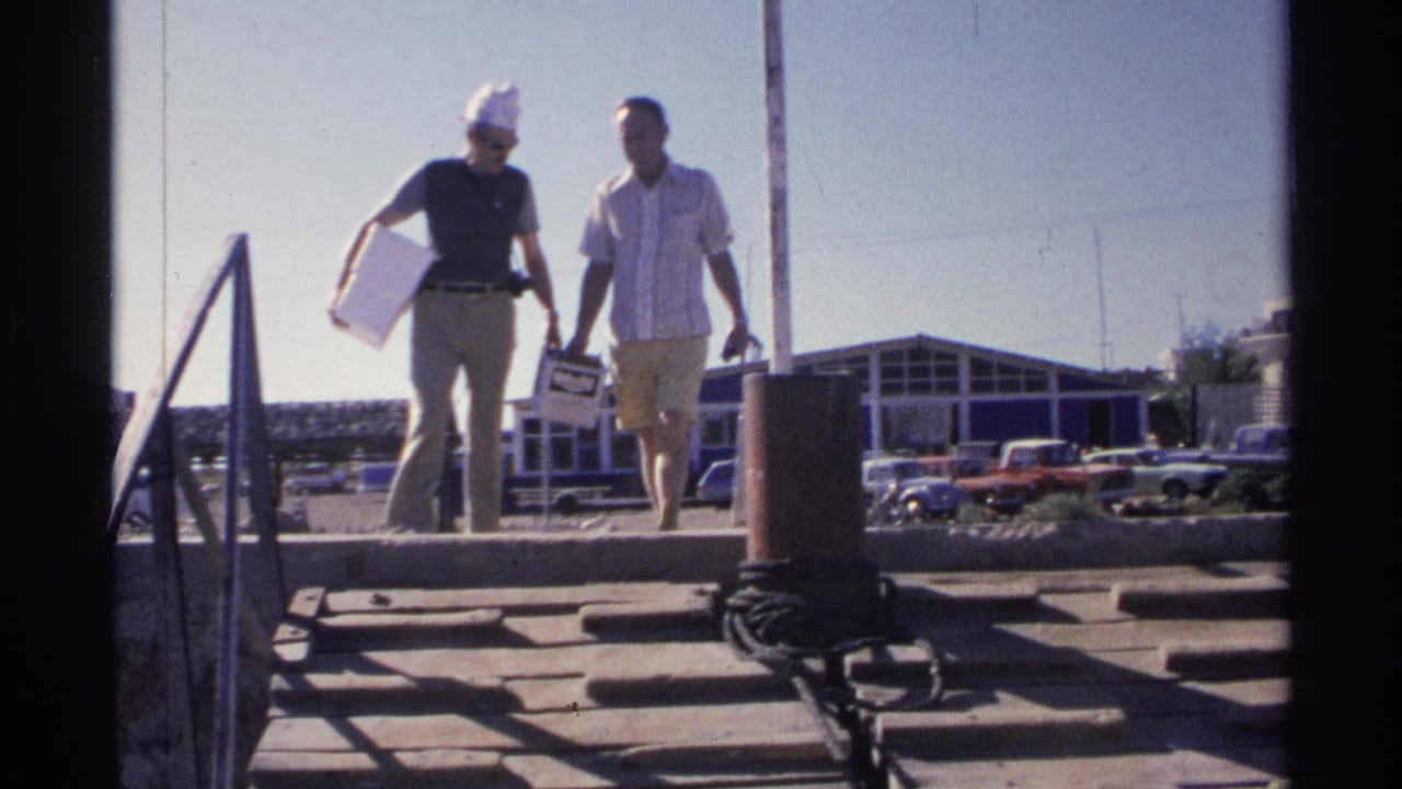 Two men walk up dock steps carrying white boxes in bright sunlight.