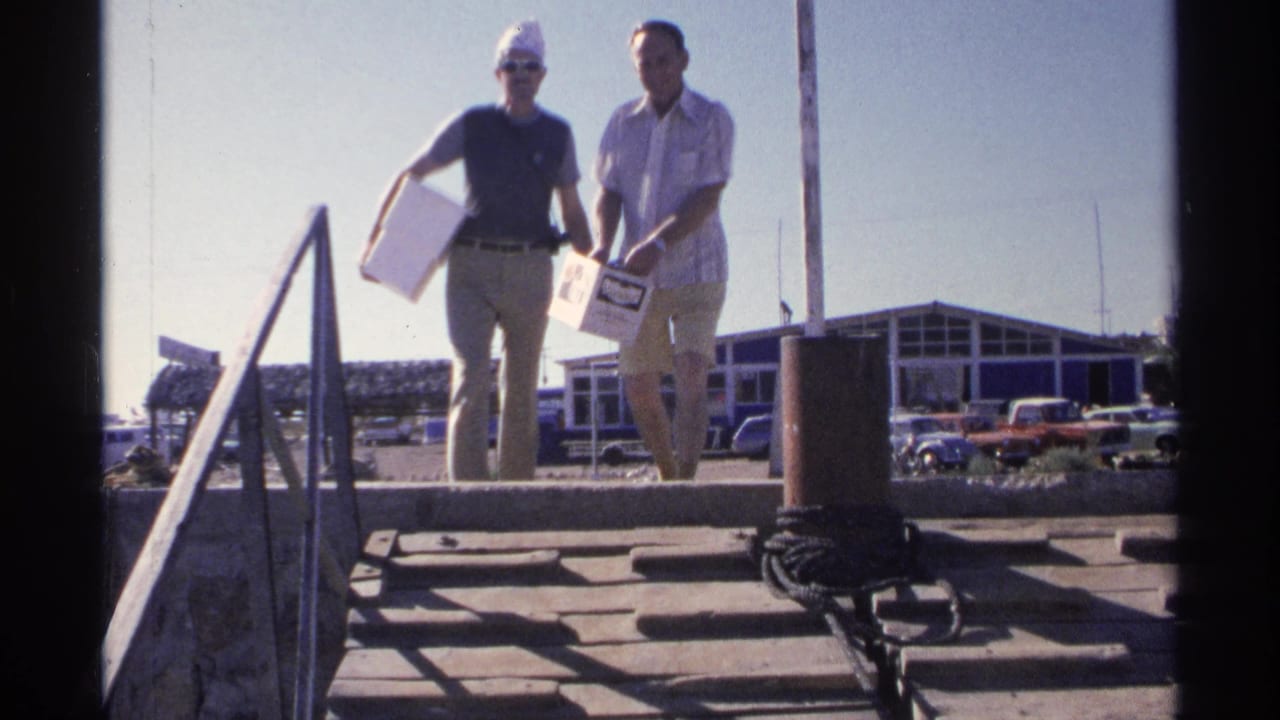 Two men stand closer on dock steps with boxes and a fish-shaped item visible.