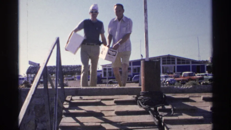 The men near the camera on a sunny Guaymas dock, one in a white hat and sunglasses and the other holding a small catch or bag.