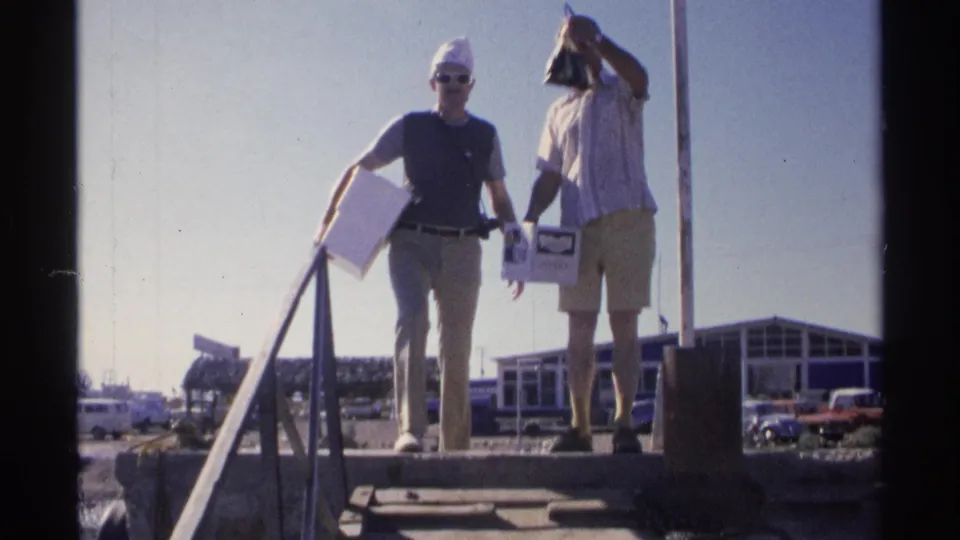 A man raises a dark fish or bag beside his companion on the dock, with a marina-side building and parked cars in the background.