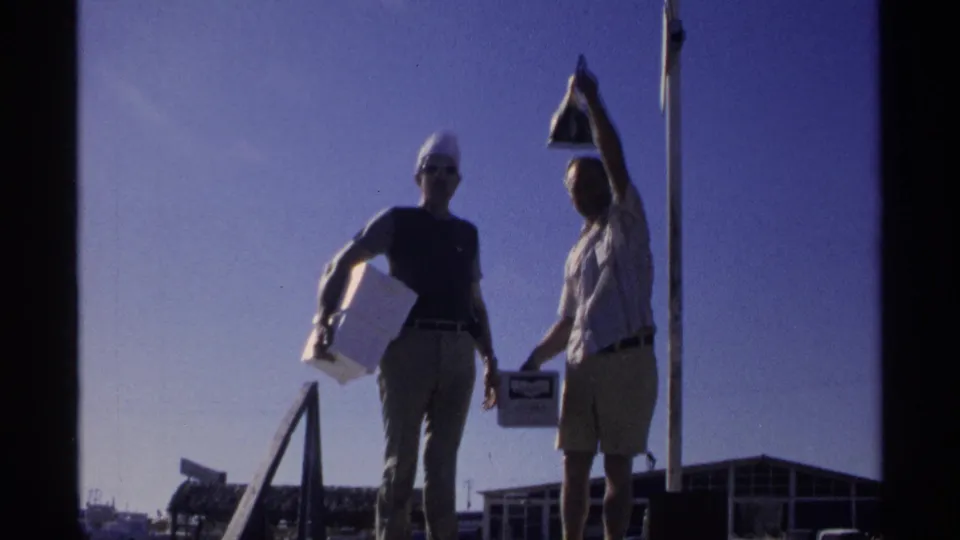 Backlit on a Guaymas dock, one man holds a dark fish or bag overhead while his companion stands with a white box.