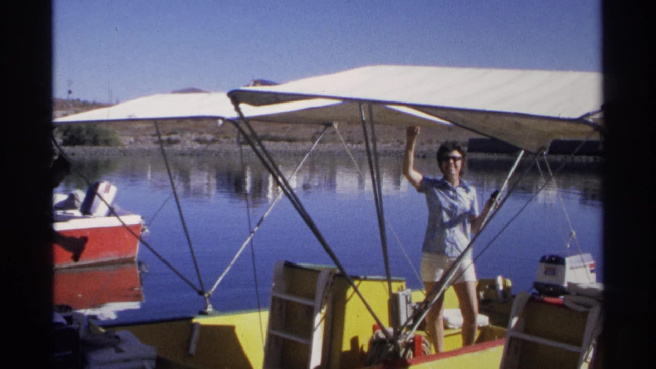 Woman standing on a yellow boat under a white canopy and waving beside calm water.