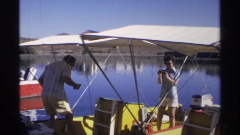 A man moves across the small boat area while the woman holds her camera under the canopy, with calm water behind them.