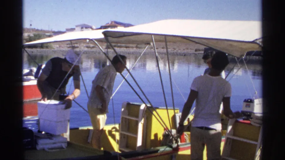 Adults in casual 1970s clothing prepare a yellow motorboat in Guaymas, Mexico, with still water and shoreline behind them.
