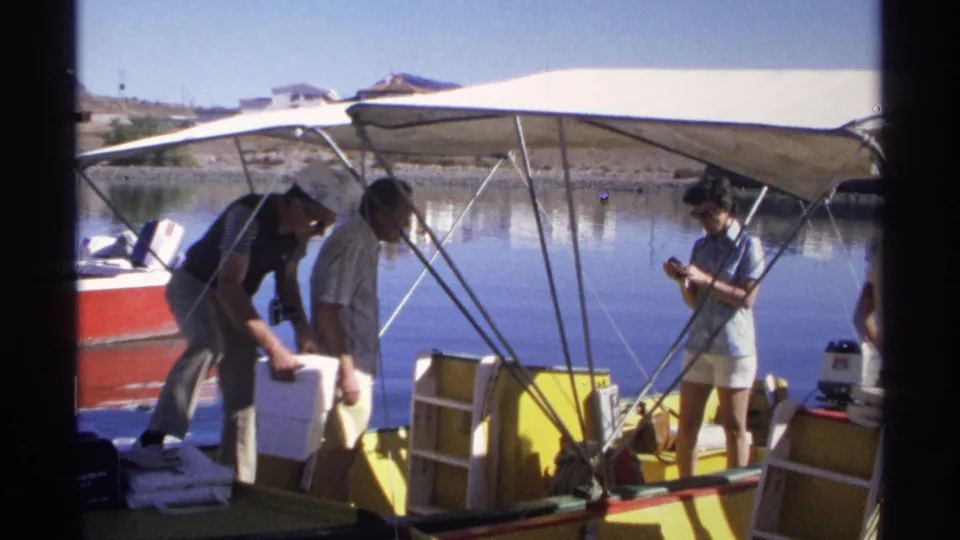 A 1975 Guaymas home-movie frame shows adults moving a cooler on a yellow motorboat while a woman holds a camera nearby.
