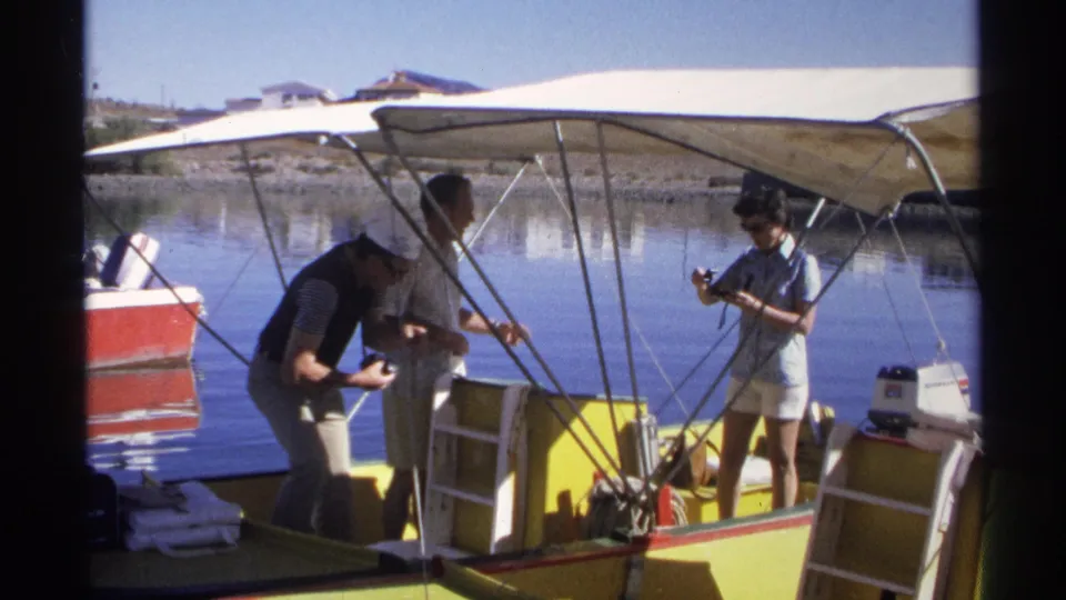 Three people in 1975 Guaymas footage handle gear on a yellow and white motorboat under a canvas canopy.