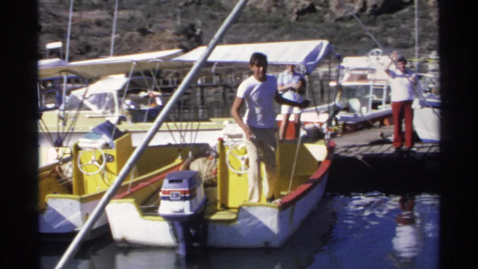 A man stands on the yellow and white motorboat near a dock in Guaymas, with other marina boats and people visible behind him.