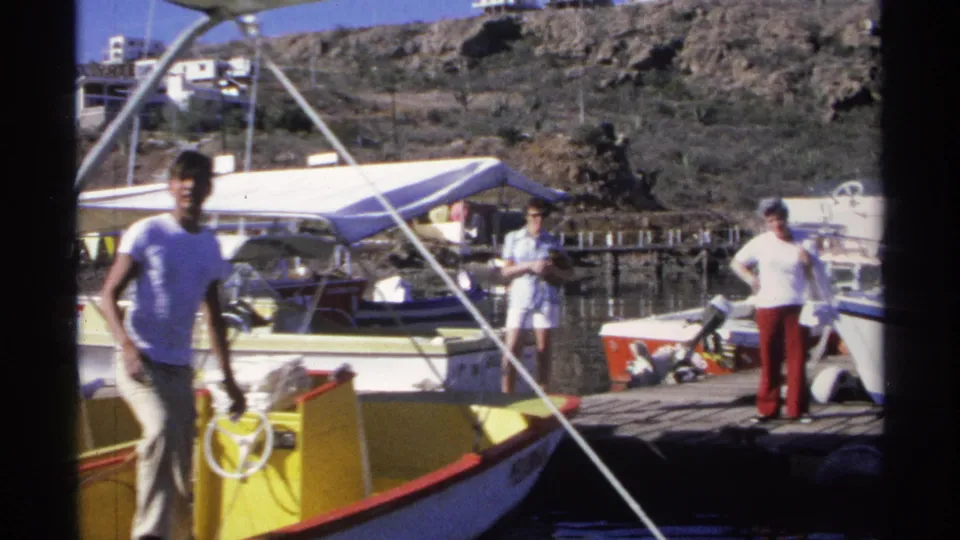 The 1975 home-movie view shows a young man moving on the yellow boat while adults stand nearby on the marina dock in Guaymas.