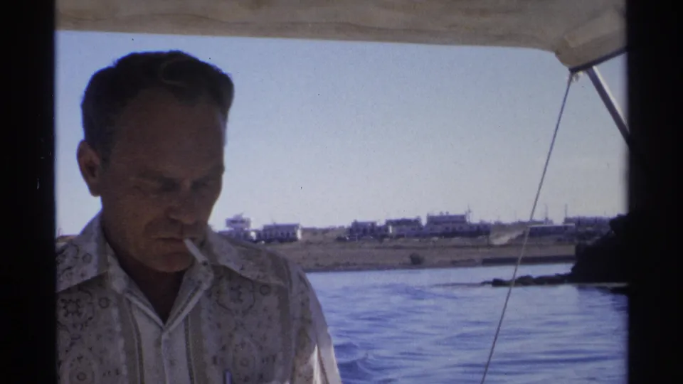 A man in a patterned shirt is filmed from close range on a boat, with Guaymas water and distant shoreline buildings behind him.