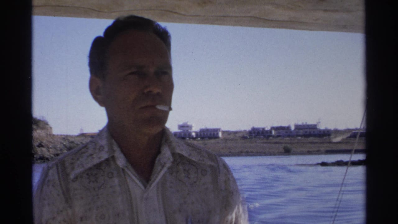 Man with a cigarette stands on a moving boat with water and shoreline behind him.